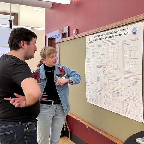 Two people stand at a poster board as one explains research presented on the poster