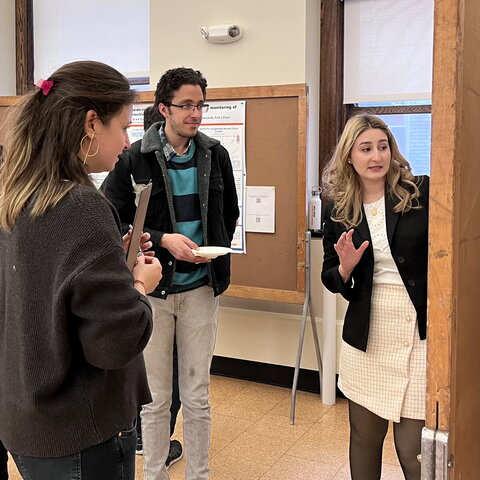 Three people stand in a classroom as one on right presents her research from a poster board.